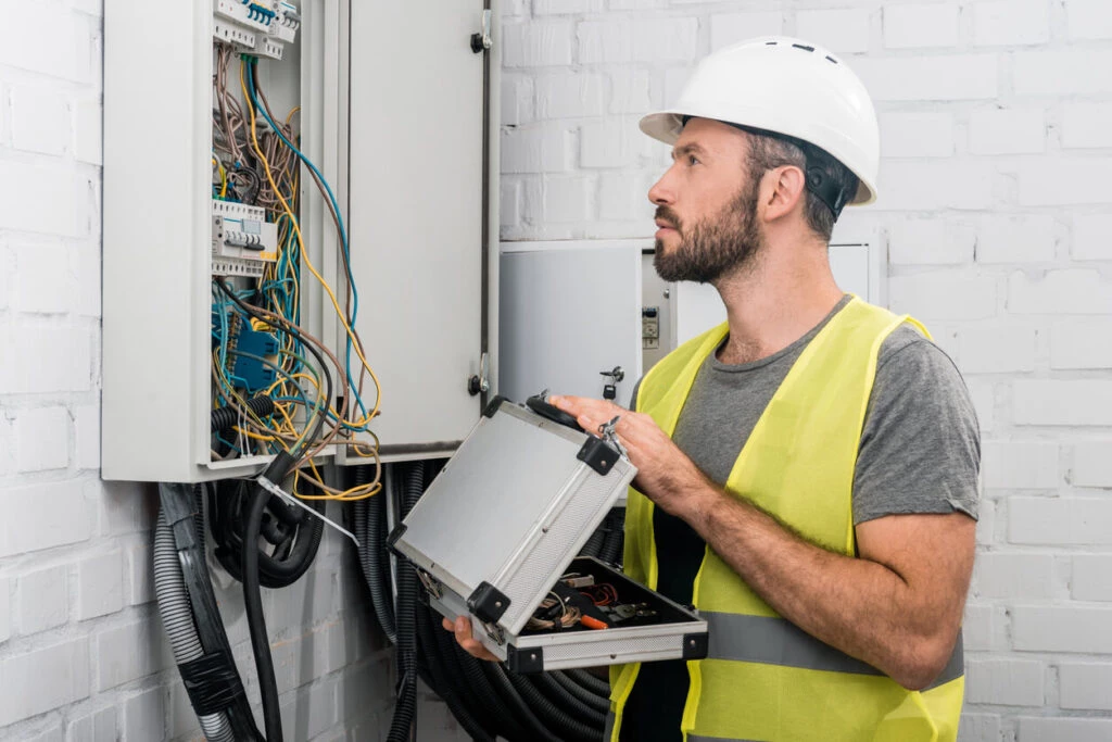 Electrician holding a toolbox and looking at an open electrical panel